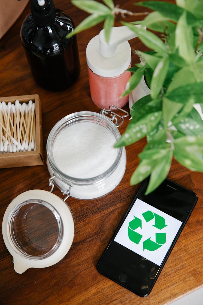 Top view of smartphone with recycling symbol on screen and eco friendly cream in glass jar, ear sticks in wooden box and soap dispensers placed on table near green leaves plant